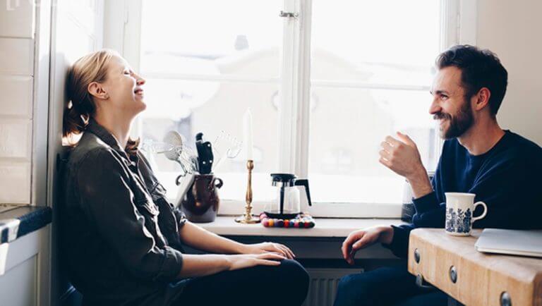 En man och en kvinna sitter vid ett köksbord, dricker kaffe och ler mot varandra.